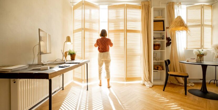Woman opens window blinds letting the sun inside the room, spending good morning in sunny and cozy apartment in beige tones. Interior view