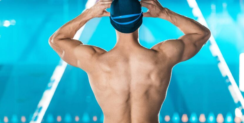 back view of muscular swimmer in swimming cap and goggles standing at swimming pool